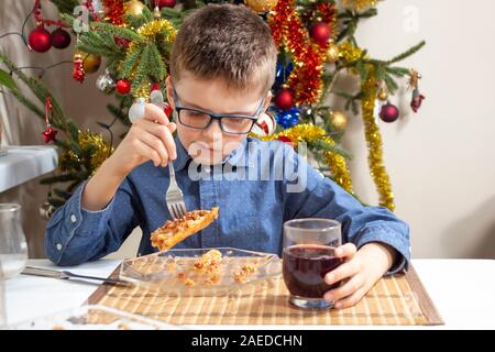 Il ragazzo con gli occhiali si siede al tavolo e il cibo di poke sulla sua forcella sulla piastra. Un bellissimo albero di Natale decorato in background. Foto Stock