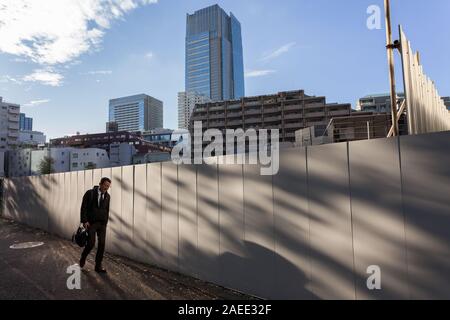 Un salarista giapponese cammina accanto a un muro di cantiere con la torre del centro di Tokyo alle spalle, Roppongi, Tokyo, Giappone. Foto Stock