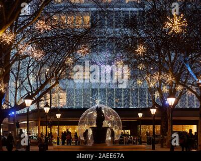 Le luci di Natale a Sloan Square. La silhouette di persone, alberi e fontana sono visti contro illuminate vetrine dei negozi al dettaglio. Foto Stock