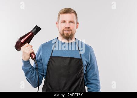 Giovani barbuto parrucchiere in grembiule blu e pullover holding asciugacapelli Foto Stock