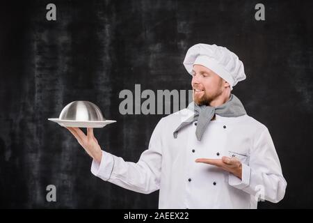 Giovani chef sorridente in uniforme cercando e che puntano a cloche con pasto cucinato Foto Stock