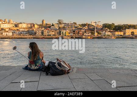 Una giovane donna con un bastone selfie guardando attraverso il fiume Douro verso Cais da Ribeira, Porto, Portogallo Foto Stock