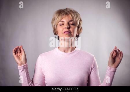 Persone, yoga e uno stile di vita sano. Splendida bionda e giovane donna vestita di luce rosa maglione mantenendo gli occhi chiusi mentre medita in ambienti chiusi, praticanti Foto Stock