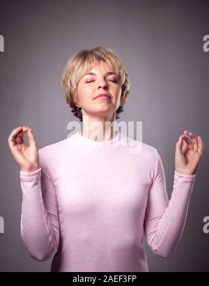 Persone, yoga e uno stile di vita sano. Splendida bionda e giovane donna vestita di luce rosa maglione mantenendo gli occhi chiusi mentre medita in ambienti chiusi, praticanti Foto Stock