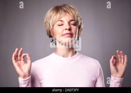 Persone, yoga e uno stile di vita sano. Splendida bionda e giovane donna vestita di luce rosa maglione mantenendo gli occhi chiusi mentre medita in ambienti chiusi, praticanti Foto Stock