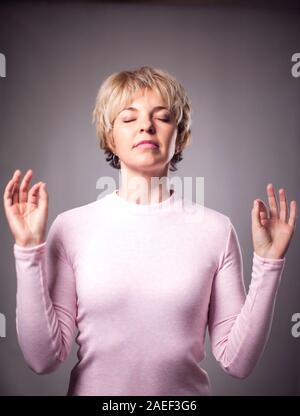 Persone, yoga e uno stile di vita sano. Splendida bionda e giovane donna vestita di luce rosa maglione mantenendo gli occhi chiusi mentre medita in ambienti chiusi, praticanti Foto Stock