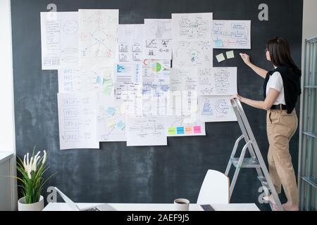 Giovane donna con un pezzo di gesso in piedi sulla scaletta nella parte anteriore di Blackboard Foto Stock