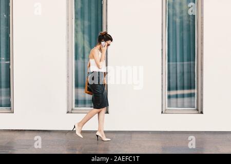 La squisita business donna in pelle gonna e camicia di seta a parlare al telefono mentre si cammina nei pressi di un edificio con grandi finestre Foto Stock