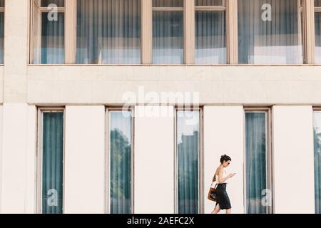 La squisita business donna in pelle gonna e camicia di seta a parlare al telefono mentre si cammina nei pressi di un edificio con grandi finestre Foto Stock