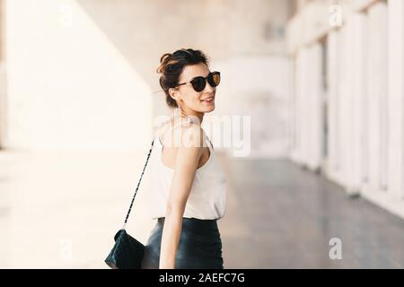 Bella sorridente giovane donna in occhiali da sole che indossa la frizione in pelle e mantello bianco facile camicetta e guardando lontano sopra la spalla Foto Stock