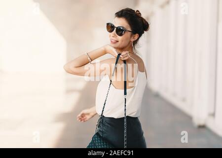 Bella sorridente giovane donna in occhiali da sole che indossa la frizione in pelle e mantello bianco facile camicetta e guardando lontano sopra la spalla Foto Stock