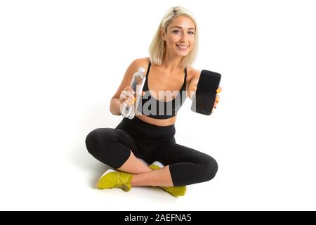 Sorridente ragazza bionda in un sport nero uniforme con giallo sneakers siede su un tappeto con una bottiglia di acqua minerale e un telefono su una bianca di sfondo per studio Foto Stock