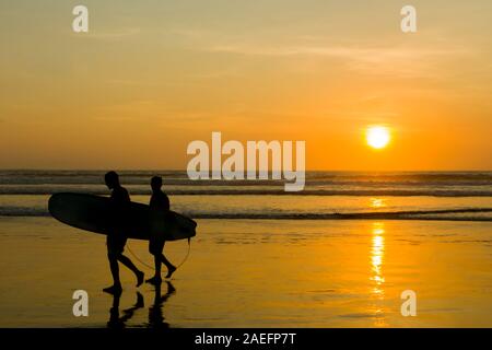 Sagome di surfisti con le loro tavole da surf sulla spiaggia di Kuta Beach a un tramonto dorato Foto Stock