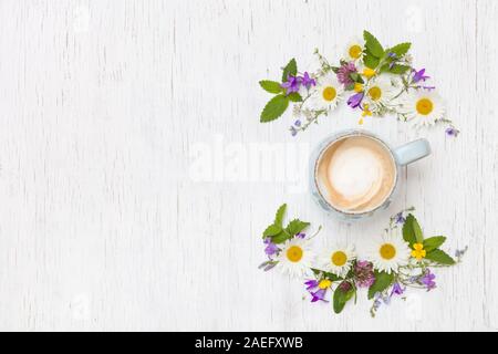 Vista dall'alto sui fiori selvatici in forma di corona e una tazza di caffè cappuccino bianco su sfondo di legno. Fiori estivi, foglie petali e co Foto Stock