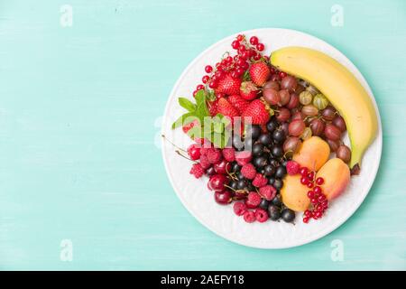 Assortimento di frutta succosa sulla piastra bianca e turchese sfondo tabella. Le banane, le fragole, mirtilli, lamponi, uva spina e ribes nero dec Foto Stock