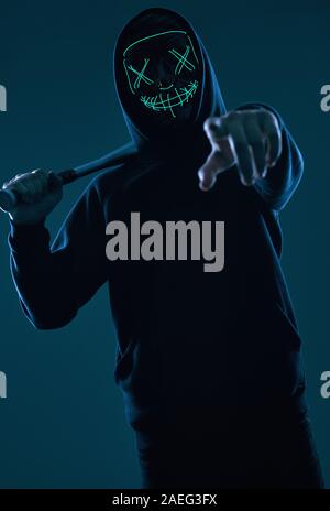 Ritratto di un anonimo uomo delinquente con la mazza da baseball in una felpa con cappuccio nero nasconde il suo volto dietro una maschera al neon. Studio shot. Foto Stock