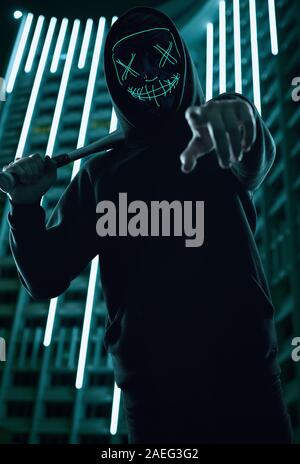 Ritratto di un anonimo uomo delinquente con la mazza da baseball in una felpa con cappuccio nero nasconde il suo volto dietro una maschera al neon. Città luminosa sullo sfondo Foto Stock