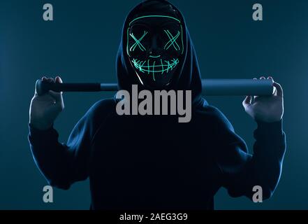 Ritratto di un anonimo uomo delinquente con la mazza da baseball in una felpa con cappuccio nero nasconde il suo volto dietro una maschera al neon. Studio shot. Foto Stock