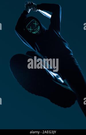 Ritratto di un anonimo uomo delinquente con la mazza da baseball in una felpa con cappuccio nero nasconde il suo volto dietro una maschera al neon. Angolo inferiore Foto Stock