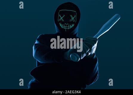 Ritratto di un anonimo uomo delinquente con la mazza da baseball in una felpa con cappuccio nero nasconde il suo volto dietro una maschera al neon. Angolo inferiore Foto Stock