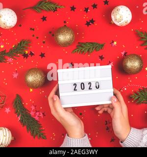 2020 è scritto su una lampada decorativa in mani femminili tra le decorazioni di Natale su uno sfondo di colore rosso. Top View layout. Foto Stock