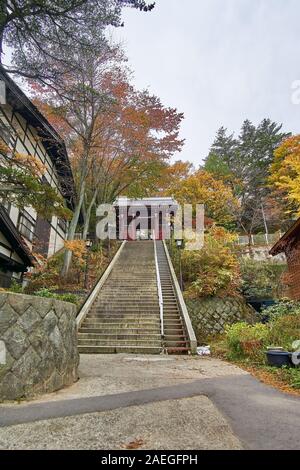 La porta rossa del Tempio di Kosenji a Kusatsu, Gunma, Giappone, si trova in cima a una scala di pietra in un giorno d'autunno, circondata da colori autunnali. Foto Stock