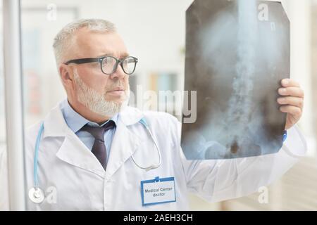 Grave chirurgo maturo indossando occhiali azienda x-ray in mano e di esame permanente, mentre in ufficio Foto Stock