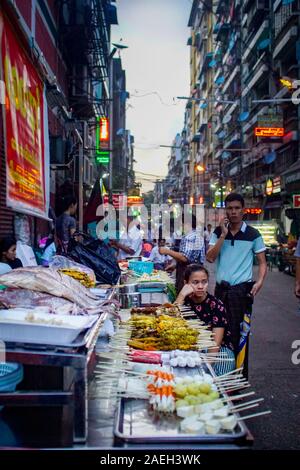 Un venditore ambulante sedersi accanto al suo bancarella vendendo gli spiedini di fresco in occasione della diciannovesima strada del mercato notturno nel quartiere di Chinatown di Yangon, Myanmar Foto Stock