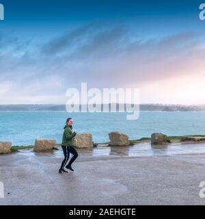 Una giovane donna sorridente e jogging lungo un vuoto costiera del parco auto in Newquay in Cornovaglia. Foto Stock