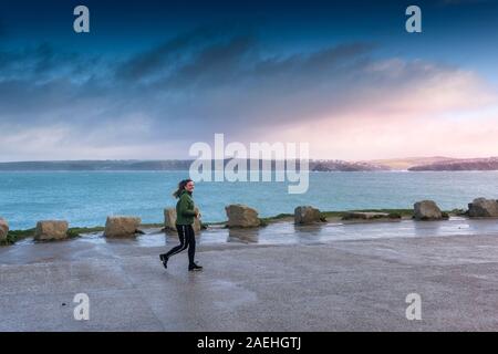 Una giovane donna sorridente e jogging lungo un vuoto costiera del parco auto in Newquay in Cornovaglia. Foto Stock
