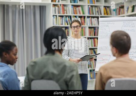 Giovane donna in occhiali in piedi vicino alla lavagna e presentando la sua relazione per la gente di affari durante una presentazione Foto Stock