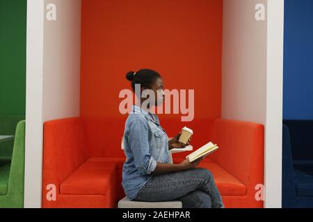 Vista laterale dello studente africano in jeans seduta di usura ed a concentrarsi sulla lettura di un libro e tenendo una tazza di caffè in mano Foto Stock