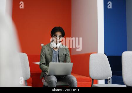 Uomo asiatico in occhiali seduti su una sedia e la digitazione sul computer portatile egli lavora online Foto Stock