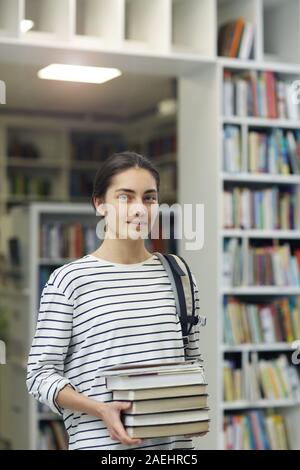 Ritratto di giovane donna bellissima azienda libri e guardando la telecamera mentre in piedi nella libreria Foto Stock
