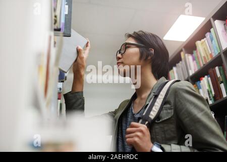 Asian giovane studente in occhiali in piedi vicino a scaffale e tenendo libro egli è nella libreria Foto Stock