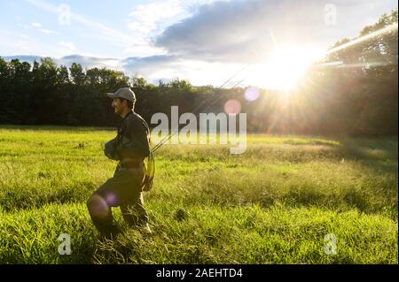 Un pescatore a mosca a piedi in un campo verso un fiume sotto il sole. Foto Stock