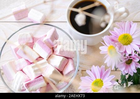 Dolce bianco rosa marshmallows in una ciotola di vetro vicino a tazza di caffè caldo e pastello crisantemo rosa fiori su una trama bianca tavolo in legno. Flatlay. Foto Stock