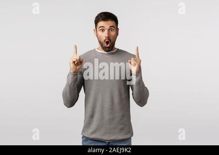 Sorpresi e stupiti e senza parole eccitato bello ragazzo barbuto in grigio maglione, dire wow, piegatura labbra divertito, affannosa stupito, dita di puntamento in alto Foto Stock