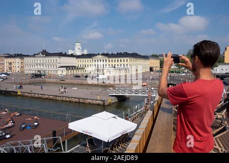Turistica prendendo foto dalla terrazza del Allas Piscina sul mare attraverso il porto del sud verso il palazzo presidenziale di Pohjoisesplanadi Street, Luthe evangelica Foto Stock