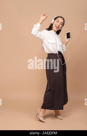 Freestyle. Giovane donna in camicia che indossano le cuffie dancing isolati su bage con lo smartphone ascoltando musica canto chiuso gli occhi felici corpo pieno Foto Stock