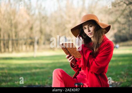 Giovane donna in total look rosso con cappotto e sciarpa leggere il libro in autunno park. La mano della bambina tocca i suoi capelli, guardando nella telecamera. Foto Stock