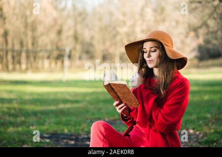 Bella bruna giovane donna in total look rosso con cappotto e sciarpa leggere il libro in autunno park. La mano della bambina tocca i suoi capelli. Foto Stock