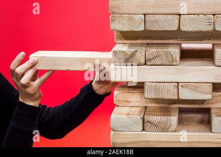 La pianificazione del rischio e della strategia nel business. La torre da blocchi di legno e la mano prendere un blocco. Gioco su sfondo rosso. Costruzione e distruzione del concetto. Il mood di pericolo e di attenzione. Foto Stock