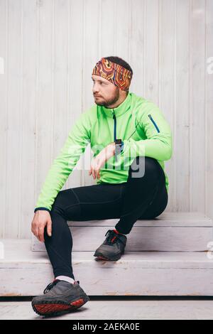 Giovane atleta di aspetto europeo in una giacca verde, guarda, pantaloni neri, si siede. Foto Stock