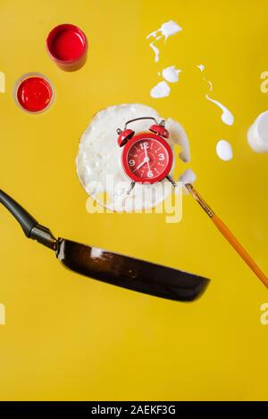 Uovo fritto e la padella con un orologio rosso, barattoli di vernice e vernici a spruzzo e a pennello levitare in aria. Foto Stock