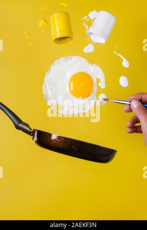 Un uovo fritto e un piattino con le lattine di vernice e un pennello salire in aria e una mano con un pennello è verniciato Foto Stock