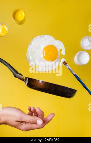 Un uovo fritto e una padella con le lattine di vernice e un pennello di vernice e di canto levitare in aria Foto Stock