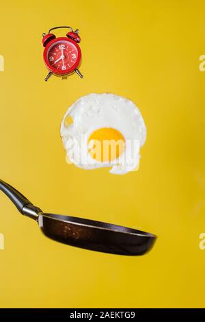 Il momento della colazione. Un uovo fritto e una padella con un orologio levitare in aria su sfondo giallo Foto Stock