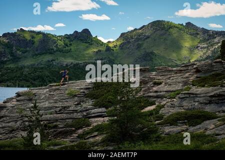 Un uomo che corre sulle rocce nel mezzo di un magnifico paesaggio di montagna. Atleta corre lungo il bordo della roccia. Uomo in maglietta blu e nero sh Foto Stock