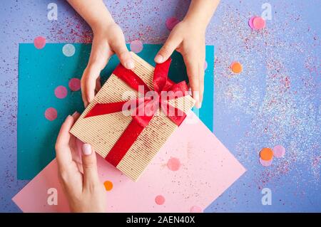 Donna e bambino le mani tenendo un giftbox legato con colore rosso vivo nastro su sfondo viola con sparklets e coriandoli. Foto Stock